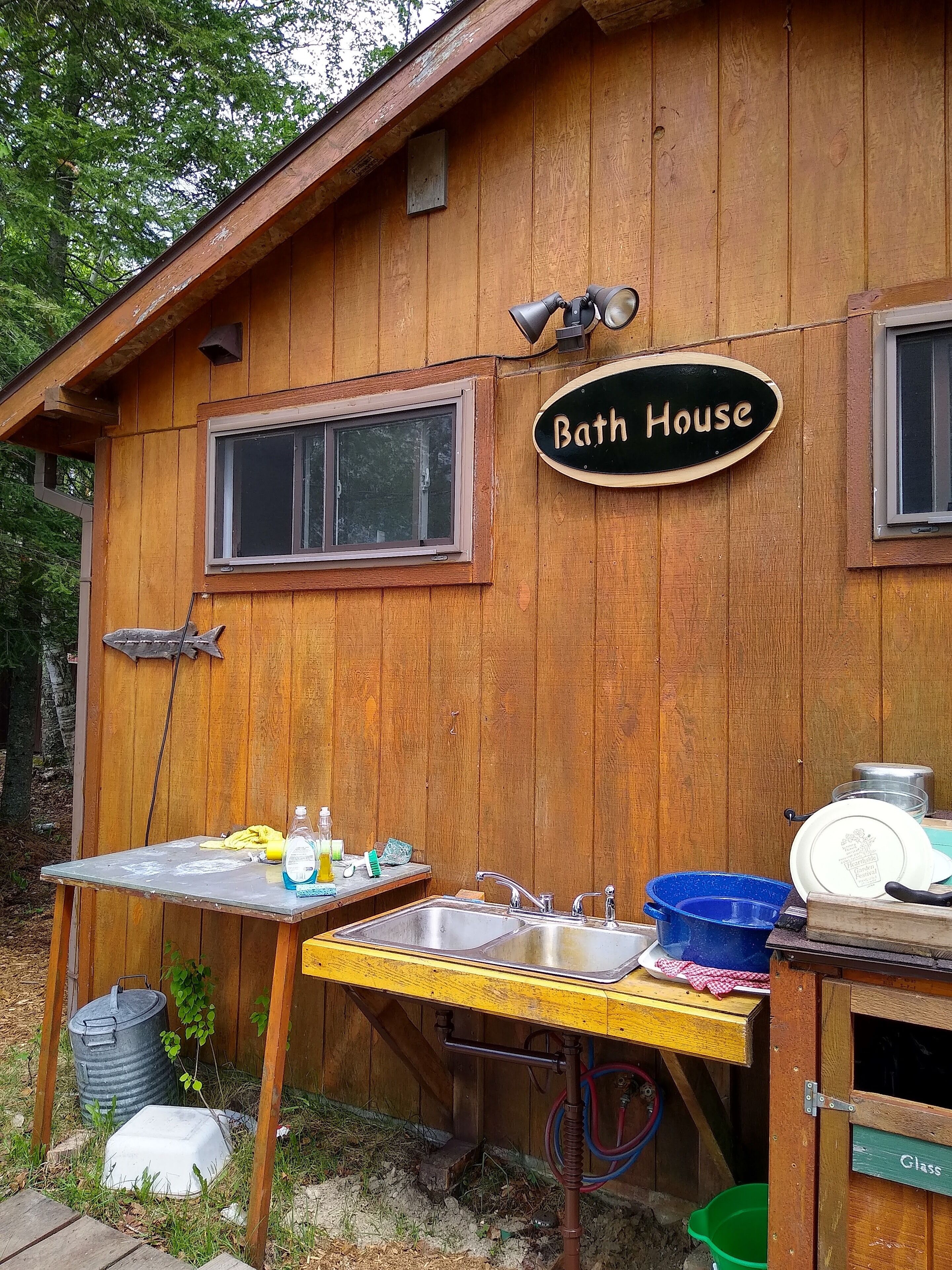 A wooden bathhouse with a sink, a table with cleaning supplies, and a sign that reads "Bath House."