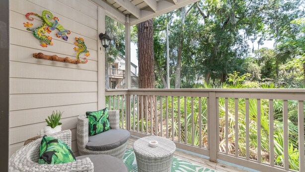 A covered porch with wicker furniture, a decorative wall piece, and a view of surrounding greenery.
