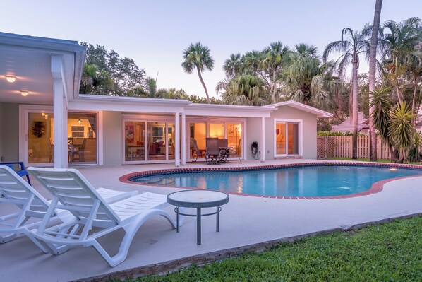 A poolside area with lounge chairs and a table, surrounded by a house with large windows and a covered patio.
