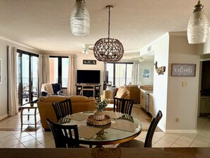 A dining area with a round glass table, chairs, a central vase, and a television in the background.