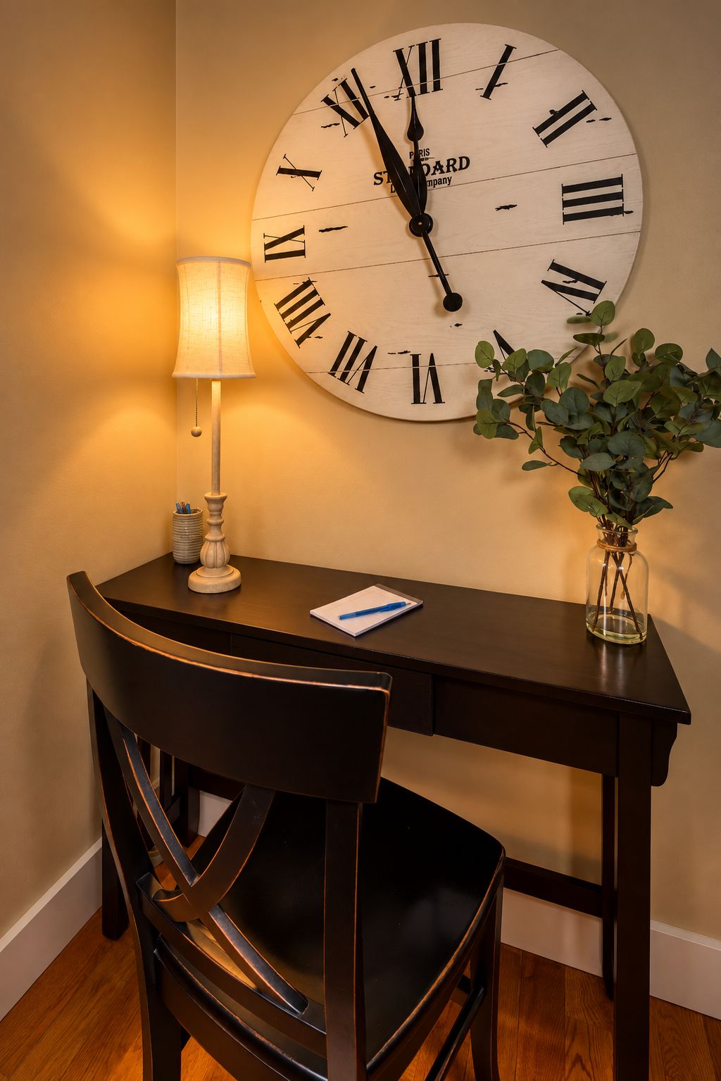 Eine Wanduhr mit römischen Ziffern, ein Schreibtisch mit Lampe, ein Stuhl, eine Vase mit Pflanze und ein Notizblock mit Stift auf dem Schreibtisch.