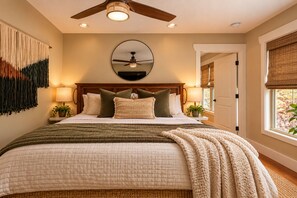 Une chambre avec un grand lit, deux lampes de chevet, un ventilateur de plafond, un miroir au-dessus du lit et une fenêtre donnant sur l’extérieur.