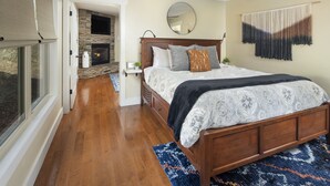 A bedroom with a large bed, a stone fireplace, a ceiling fan, and a view through an open door. - Family-Run Cottage with Fireplace and Private Hot Tub (Asheville)