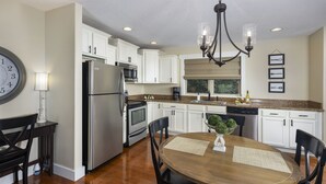 A modern kitchen with white cabinets, stainless steel appliances, and a wooden dining table. - Family-Run Cottage with Fireplace and Private Hot Tub (Asheville)
