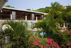 A two-story building with a balcony, surrounded by lush greenery and pink flowers.