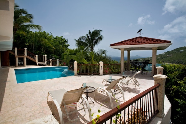 A pool area with lounge chairs and a gazebo, surrounded by greenery and a view of the landscape.