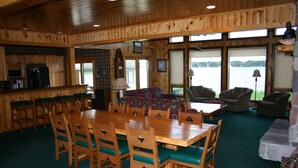 A wooden interior with a dining area, kitchen, and seating by large windows overlooking a body of water.