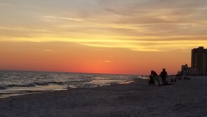 Une plage au coucher du soleil, avec des personnes qui se promènent et un bâtiment en arrière-plan.