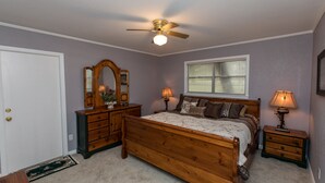 Un dormitorio con cama de madera, ventilador de techo, dos mesitas de noche con lámparas, cómoda, espejo y ventana con persianas.