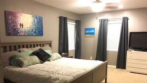 Un dormitorio con cama, mesita de noche, cómoda, ventilador de techo y dos ventanas con cortinas.