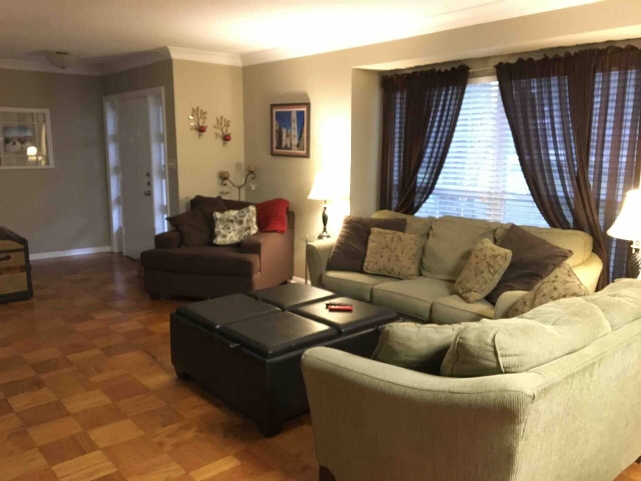 A living room with a sofa, armchair, and ottoman, a wooden floor, and a window with curtains.