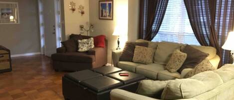 A living room with a sofa, armchair, and ottoman, a wooden floor, and a window with curtains.