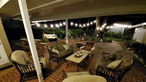 Un área de patio al aire libre con suelo de ladrillo, mobiliario de mimbre y luces colgantes.