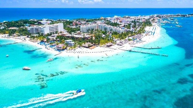 Oversigtsbillede af en kystresort med strand, krystalklart turkis vand og en mole, der strækker sig ud i havet.