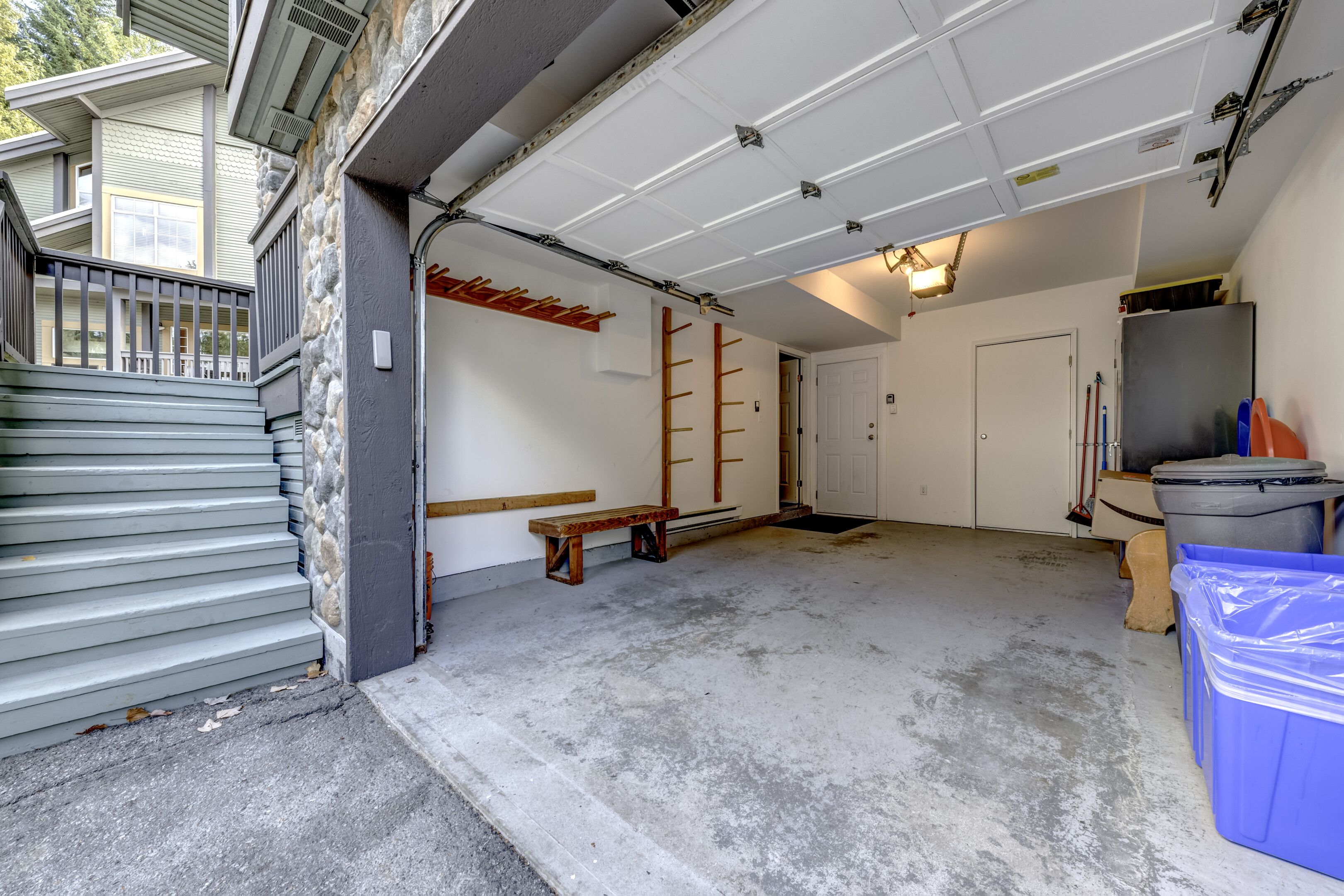 A garage with a concrete floor, a wooden bench, a door, and a trash bin.