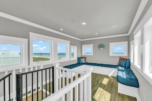A spacious room with a large window overlooking the beach, a built-in bench with cushions, and a white railing. - Top-Rated Beachfront • Steps to Sand • Hot Tub • Elevator • Firepit • Sleeps 16 (Galveston)