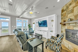 A spacious living room with a stone fireplace, a large flat-screen TV, and a view of the ocean through large windows. - Top-Rated Beachfront • Steps to Sand • Hot Tub • Elevator • Firepit • Sleeps 16 (Galveston)