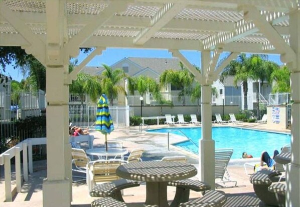 Un área de piscina techada con sillones reclinables y mesas, piscina y vista de edificios residenciales al fondo.