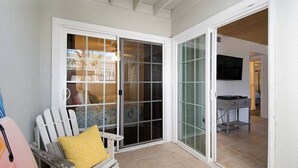 Une pièce dotée d’une porte-fenêtre coulissante donnant sur une terrasse, d’une chaise blanche avec un coussin jaune et d’un téléviseur fixé au mur.