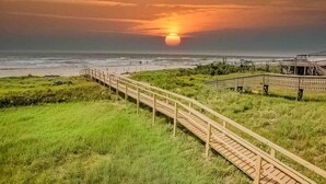 Ein hölzerner Steg führt zu einem Strand, im Hintergrund ist ein Sonnenuntergang zu sehen.