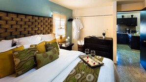 Une chambre d’hôtel avec une tête de lit ornée d’un motif en briques, un lit avec une literie verte et blanche, une table de chevet avec une lampe, une commode et un plateau avec des verres à vin.