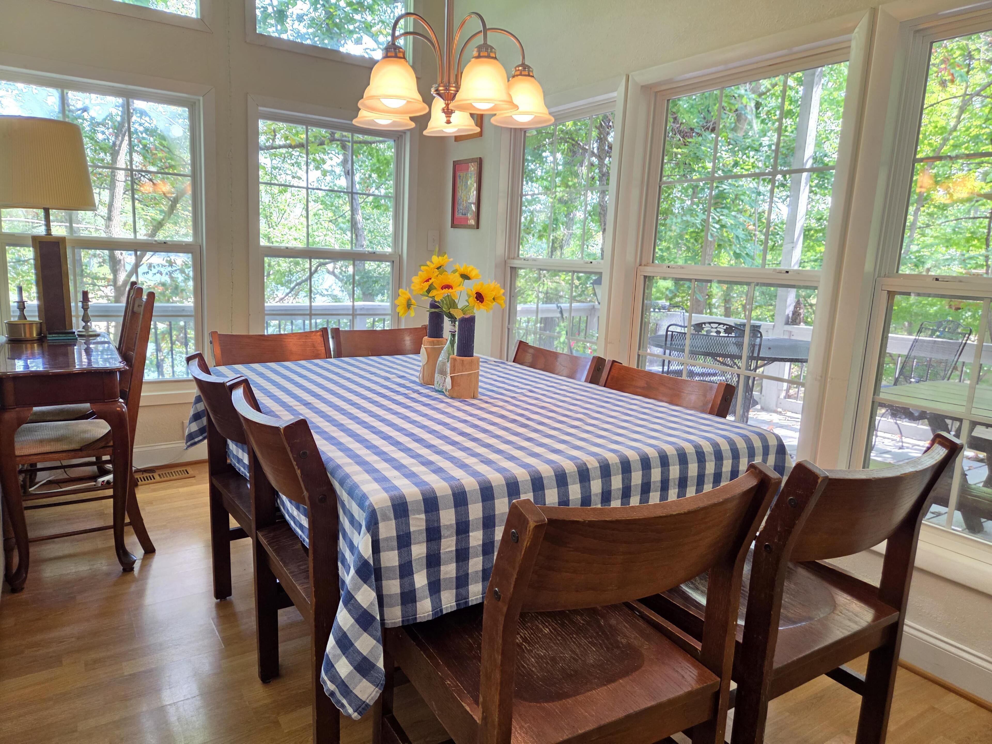 A dining room with a checkered tablecloth, wooden chairs, a vase with flowers, and large windows with a view of trees.