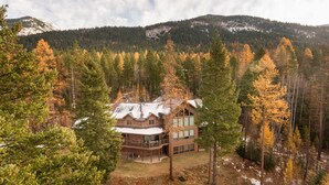 En hytte med træfacade, store vinduer og en balkon, omgivet af skov og bjerge.