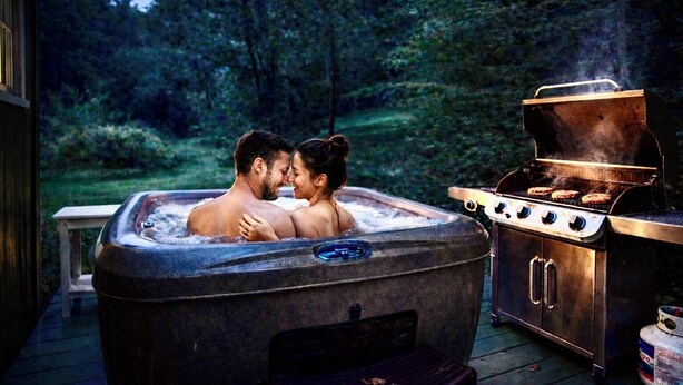 Un jacuzzi con dos personas, una parrilla con comida cocinándose y un fondo de árboles.