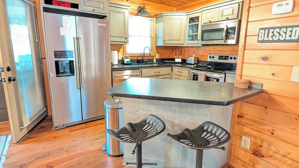 A wooden cabin interior with a kitchen featuring a refrigerator, microwave, and stove. There are two stools at a bar counter. - Whispering Creek Cabin – Peaceful, Hot Tub, Firepit & Mountain Escape (Sautee Nacoochee)