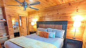 A wooden cabin bedroom with a bed, ceiling fan, bedside lamps, and a nightstand. - Whispering Creek Cabin – Peaceful, Hot Tub, Firepit & Mountain Escape (Sautee Nacoochee)