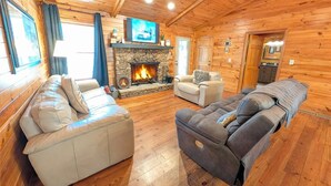 A cozy log cabin interior with a stone fireplace, leather sofas, and a television mounted above the fireplace. - Whispering Creek Cabin – Peaceful, Hot Tub, Firepit & Mountain Escape (Sautee Nacoochee)