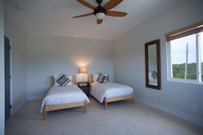 A bedroom with two single beds, a ceiling fan, a bedside table with a lamp, a mirror, and a window with a view of greenery.
