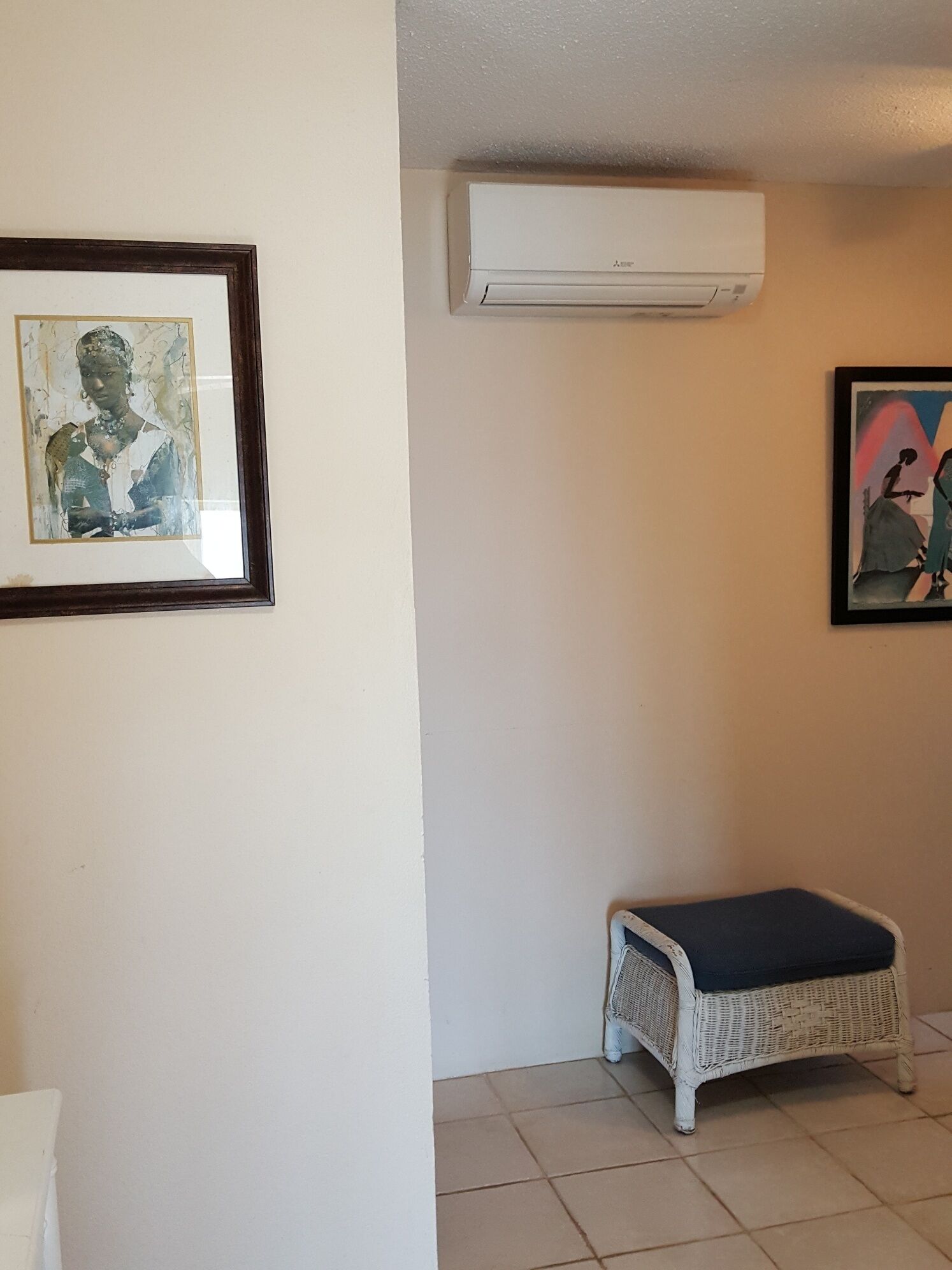 A room with a white air conditioning unit, a framed abstract painting, a wicker bench, and a framed picture on the wall.