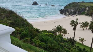 A beach view with a white balcony railing, lush greenery, and a sandy beach. - Panoramic Private Beachfront Home with Breathtaking Views (Southampton)