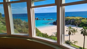A view from a window overlooking a beach with clear blue water, rocks, and palm trees. - Panoramic Private Beachfront Home with Breathtaking Views (Southampton)