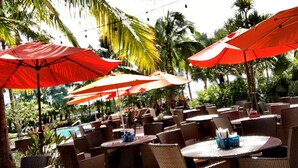 Espace extérieur aménagé avec des chaises et des tables en osier, des parasols rouges et une piscine en arrière-plan.
