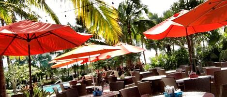 Outdoor seating with wicker chairs and tables, red umbrellas, and a pool in the background.