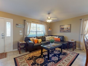 A living room with a leather sofa, a coffee table, a patterned rug, a ceiling fan, and a window with curtains.