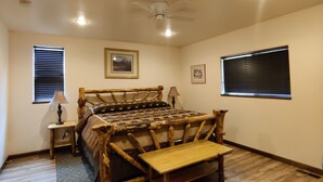 Ein Schlafzimmer mit einem hölzernen Bett, Deckenventilator, zwei Fenstern mit Jalousien und einem kleinen Nachttisch mit Lampe.