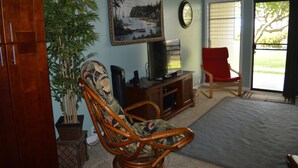 A living room with a wooden chair, a television on a wooden stand, a potted plant, and a large window with blinds. - Peaceful Ocean View. Ground Floor. Classy, clean and quiet. (Kaunakakai)