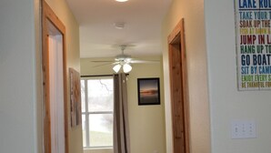 A hallway with wooden floors, white walls, and a ceiling fan. There is a window with curtains and a framed picture on the wall.