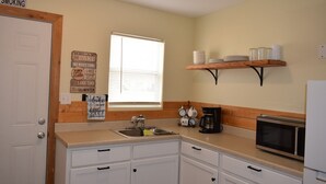 A small kitchen with white cabinets, a sink, a microwave, and a coffee maker. There is a window with blinds and a shelf above the counter holding plates and cups.