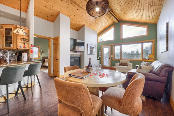 A spacious living area with a wooden ceiling, a dining table with chairs, a sofa, and a kitchen area with wooden cabinets and a television. - Pikes Peak Ranch - Bear Den Cabin (Woodland Park)
