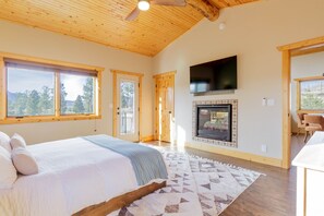 A bedroom with a large bed, a fireplace, a TV, and a view of trees through the windows. - Pikes Peak Ranch - Bear Den Cabin (Woodland Park)