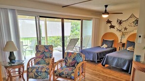 Une chambre avec deux lits, un plancher en bois, une table en verre, deux chaises recouvertes d’un tissu à motifs floraux, une lampe et un ventilateur de plafond.