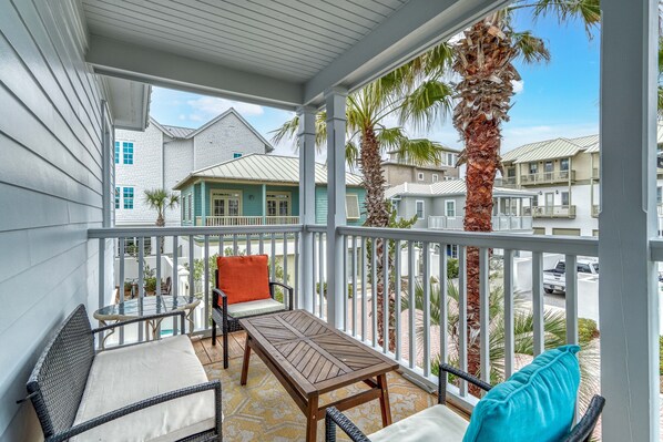 A covered balcony with wicker furniture, a wooden table, and a view of neighboring houses and palm trees.