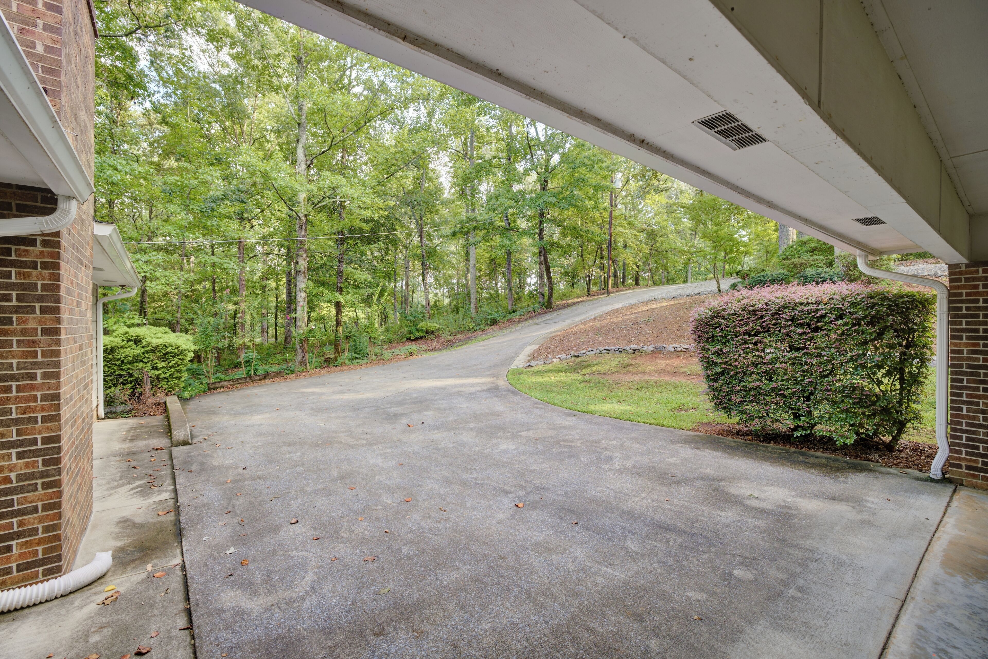 A covered driveway leading to a house with a brick exterior, surrounded by a wooded area.