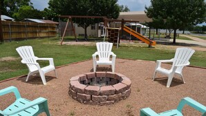 A playground with a swing set, slide, and fire pit with chairs around it. - Cabin at Texas Hills RV Haven on Lake Buchanan (Tow)