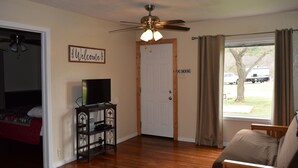 A room with a bed, a TV on a stand, a ceiling fan, a door with a 'NO SMOKING' sign, and a window with curtains. - Cabin at Texas Hills RV Haven on Lake Buchanan (Tow)