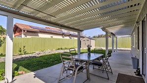 Un patio cubierto con una mesa y sillas, un jardín pequeño y una cerca verde al fondo.